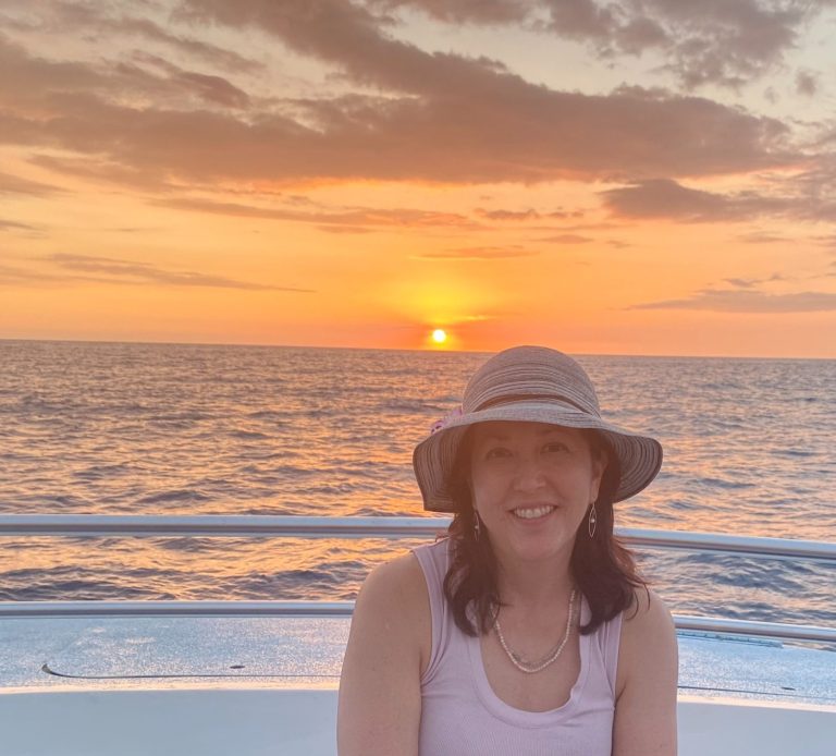 Joyce in foreground, sitting on boat, with sun setting on the ocean horizon behind her.